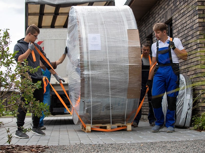 Čtyři dospělé osoby manipulují se zabaleným koupacím sudem Gardenvity na dřevěné paletě.